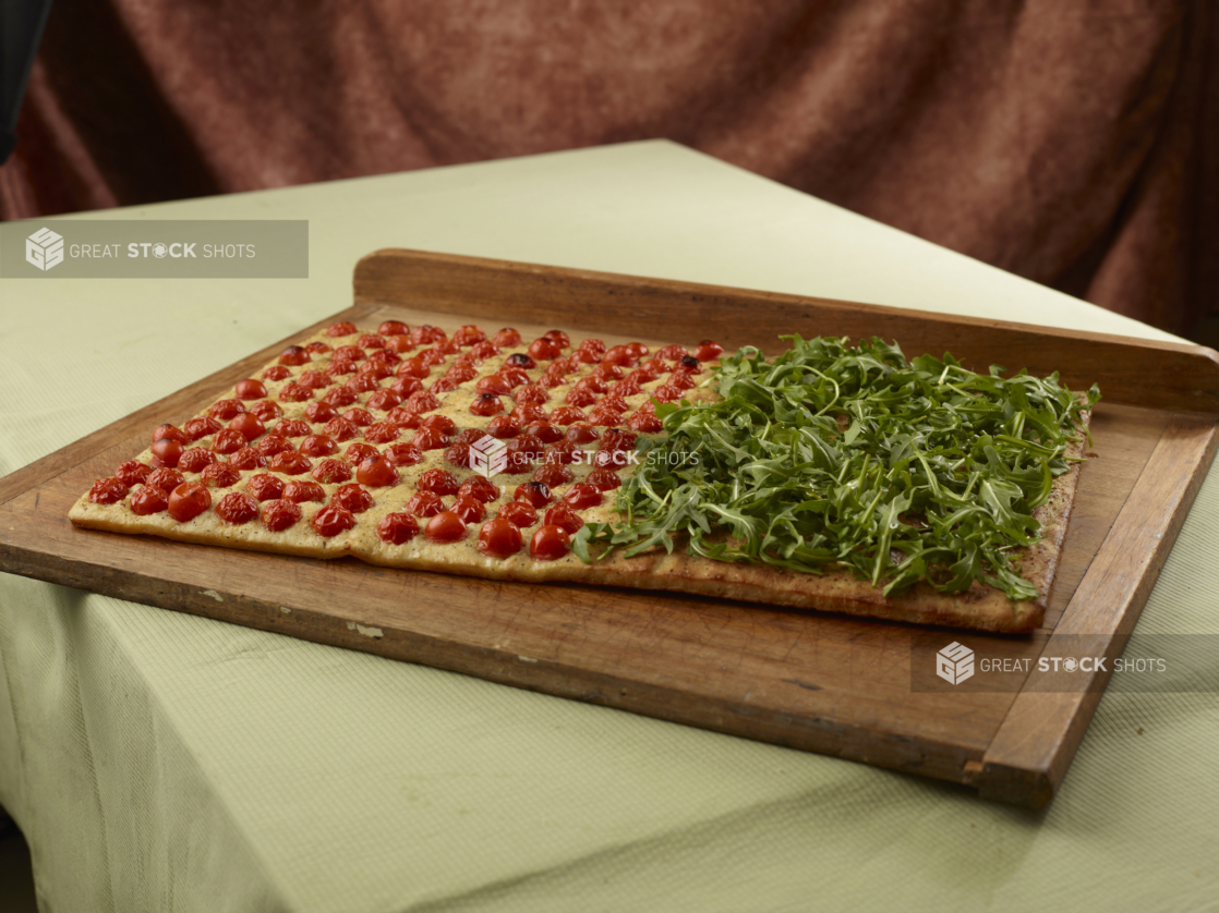 Large rectangular focaccia with arugula on half and blistered cherry tomatoes on the other on a wood board, tablecloth