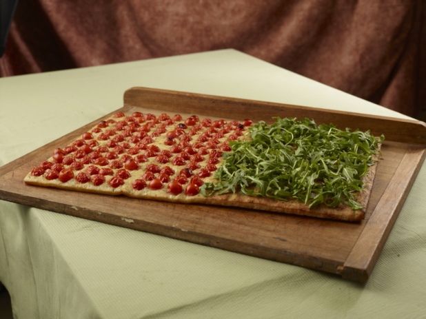 Large rectangular focaccia with arugula on half and blistered cherry tomatoes on the other on a wood board, tablecloth