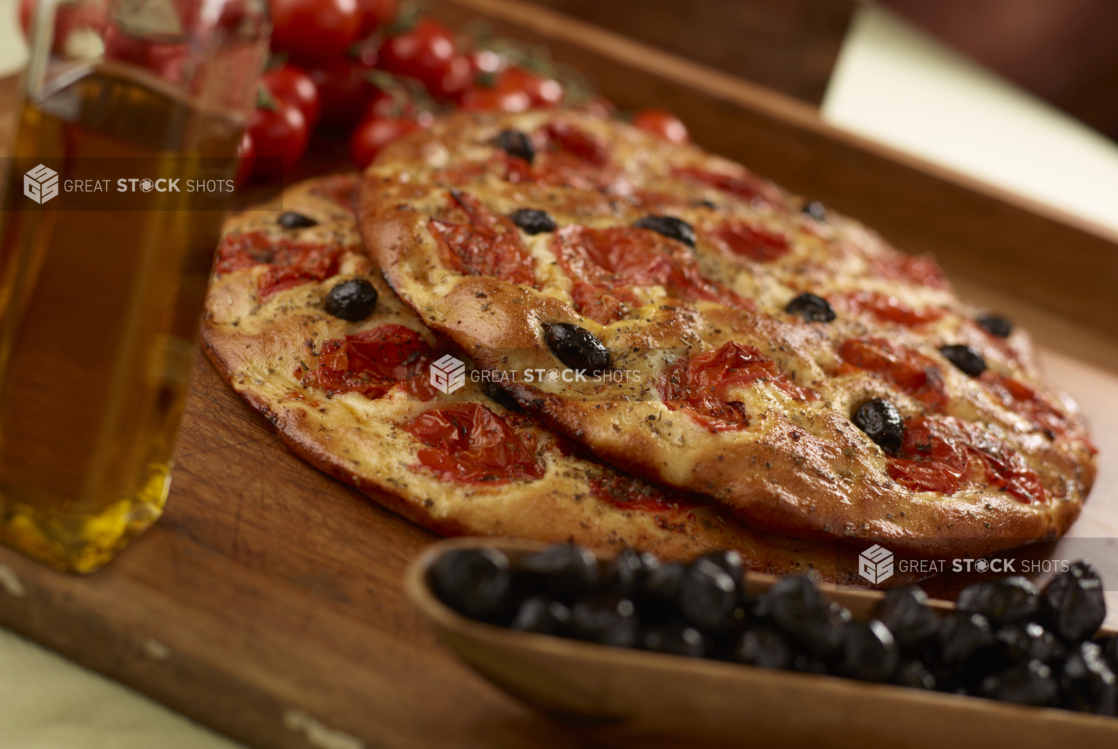 Two round herbed cherry tomato and black olive flatbreads piled on a wooden cutting board, black olives and olive oil in foreground, close-up