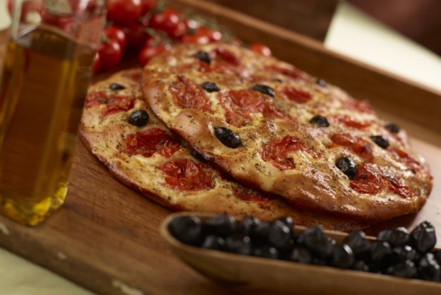 Two round herbed cherry tomato and black olive flatbreads piled on a wooden cutting board, black olives and olive oil in foreground, close-up