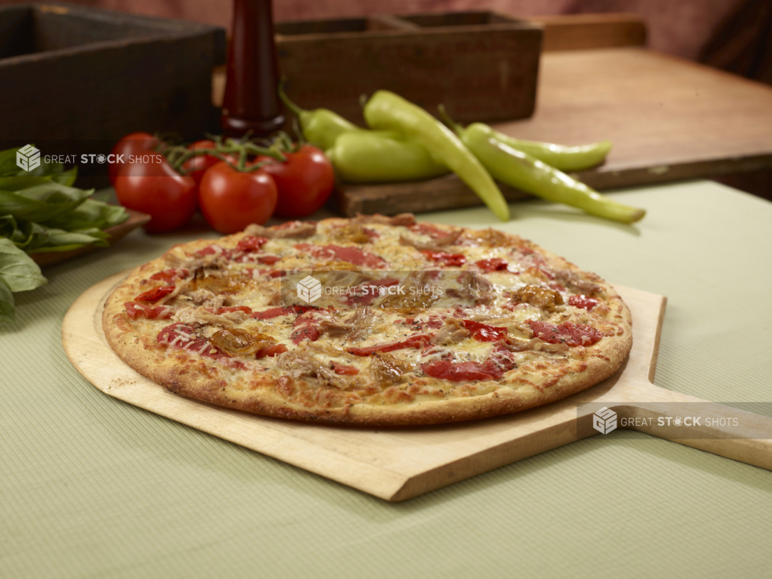 Whole unsliced pizza with pulled pork, roasted red peppers, and roasted garlic on a wood pizza peel with fresh vegetables in background, close-up, low angle