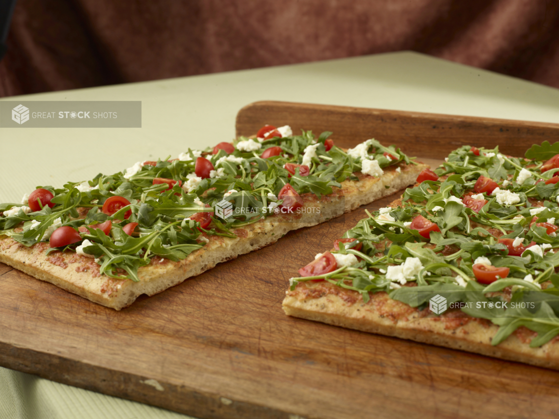 Close Up of a Cut Sicilian-Style Rectangular Pizza with Fresh Arugula, Tomato and Feta Toppings on a Wooden Board