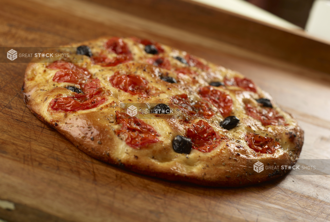 Round herbed cherry tomato and black olive flatbread on a wooden cutting board, close-up