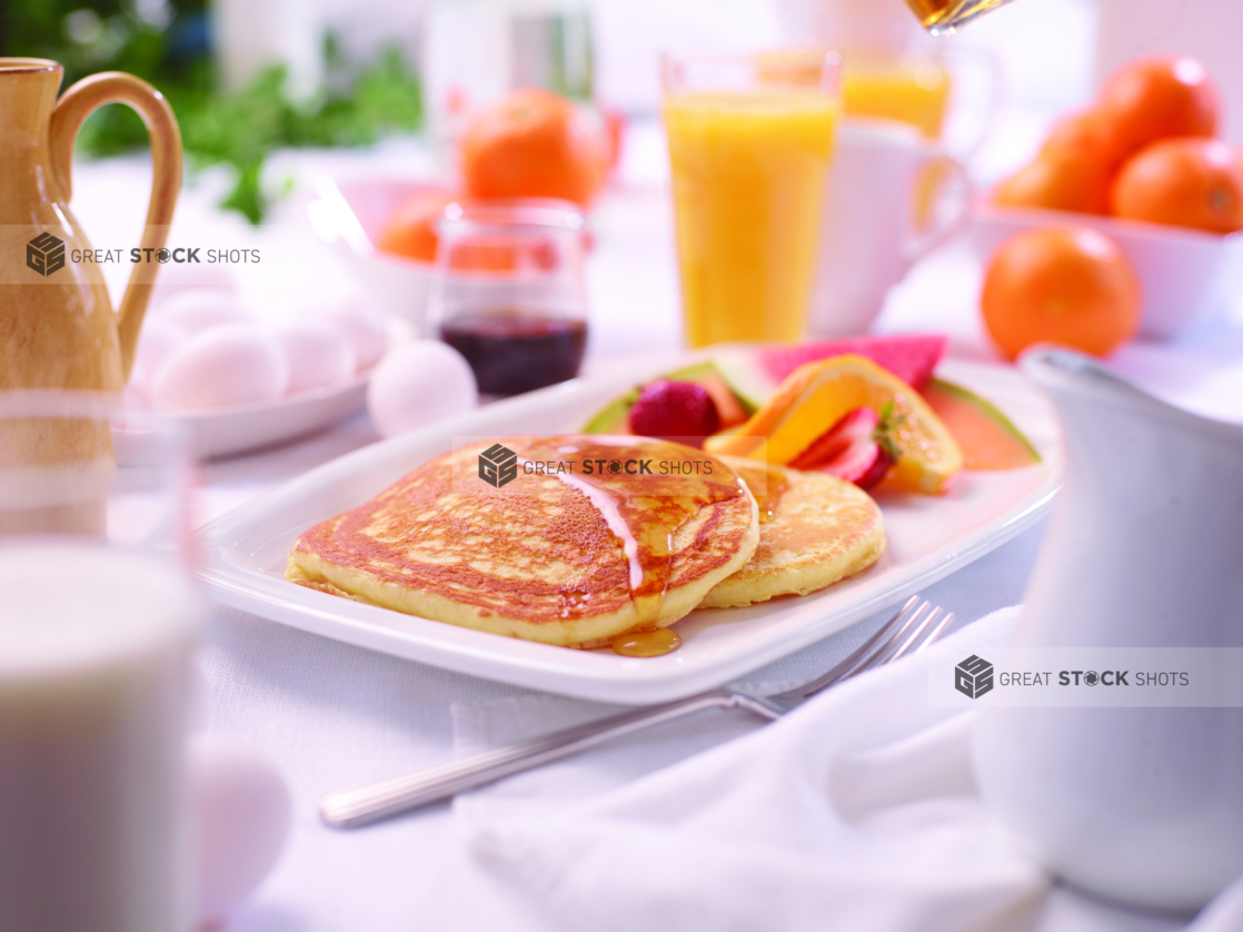 A Platter of Buttermilk Pancakes with Maple Syrup and Fresh Fruits on a White Dish in an Indoor Setting