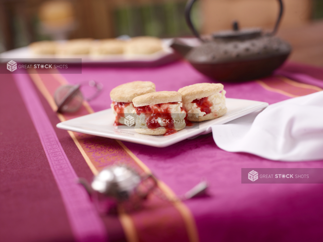 a close up of food on a aA Plate of Scone, Clotted Cream and Strawberry Jam Sandwiches on a Pink and Red Table Cloth Surfacetable