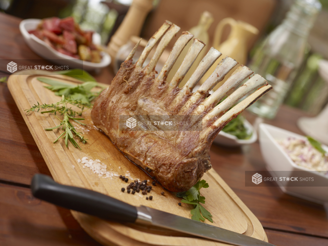 Whole rack of lamb on a wood cutting board ready for carving, buffet items in background