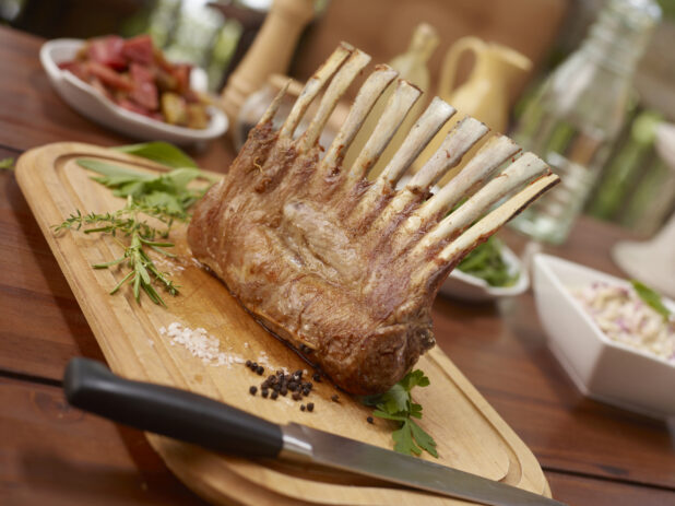 Whole rack of lamb on a wood cutting board ready for carving, buffet items in background