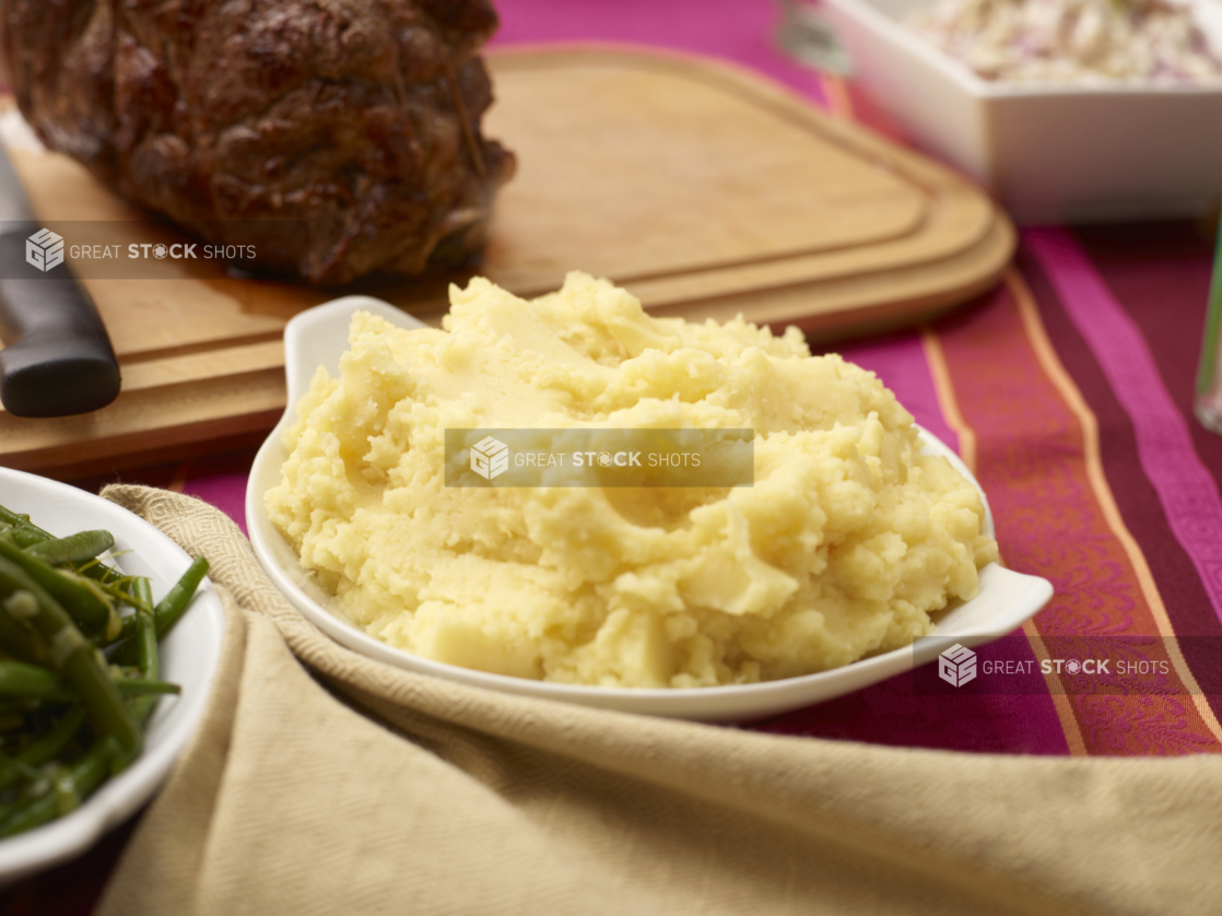 Mashed potatoes on a white platter on a buffet table with a pink tablecloth, roast meat in background