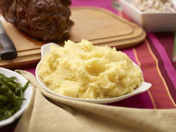 Mashed potatoes on a white platter on a buffet table with a pink tablecloth, roast meat in background