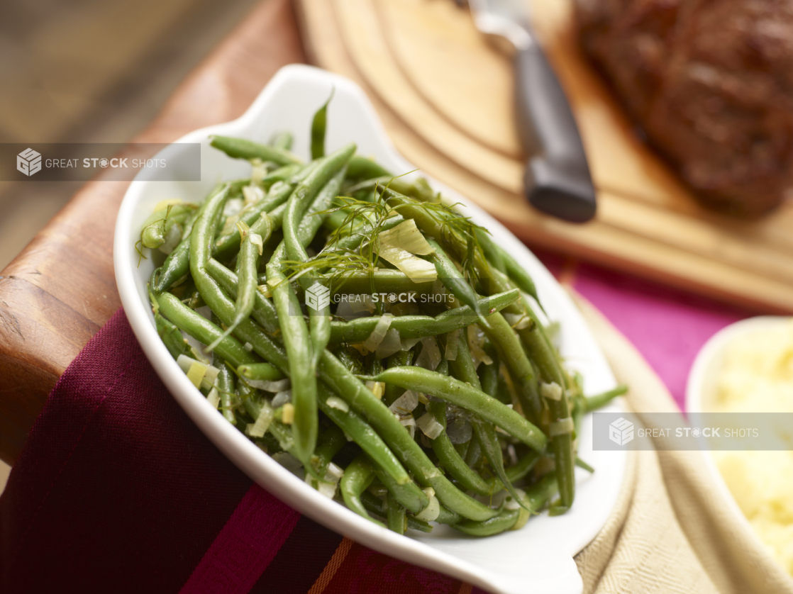 Green beans with leeks and fennel in a white serving dish, roast and mashed potatoes in background