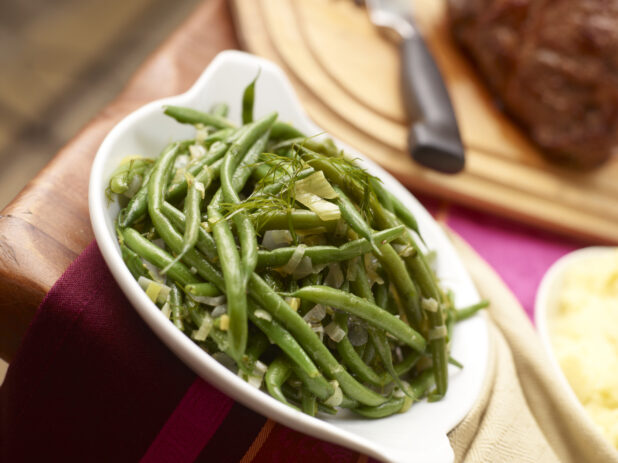 Green beans with leeks and fennel in a white serving dish, roast and mashed potatoes in background