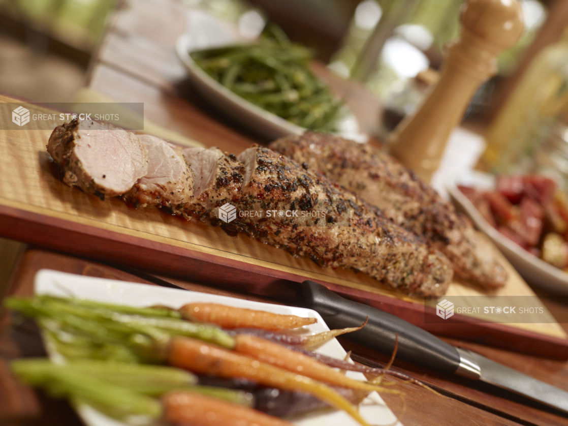 Pork tenderloin, partially sliced on a wood cutting board with side dishes surrounding