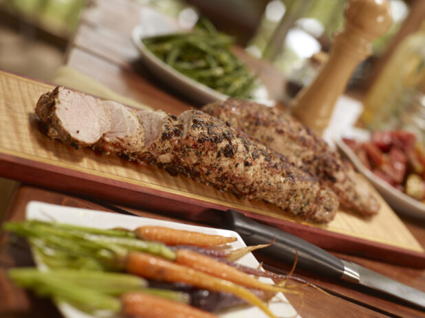 Pork tenderloin, partially sliced on a wood cutting board with side dishes surrounding