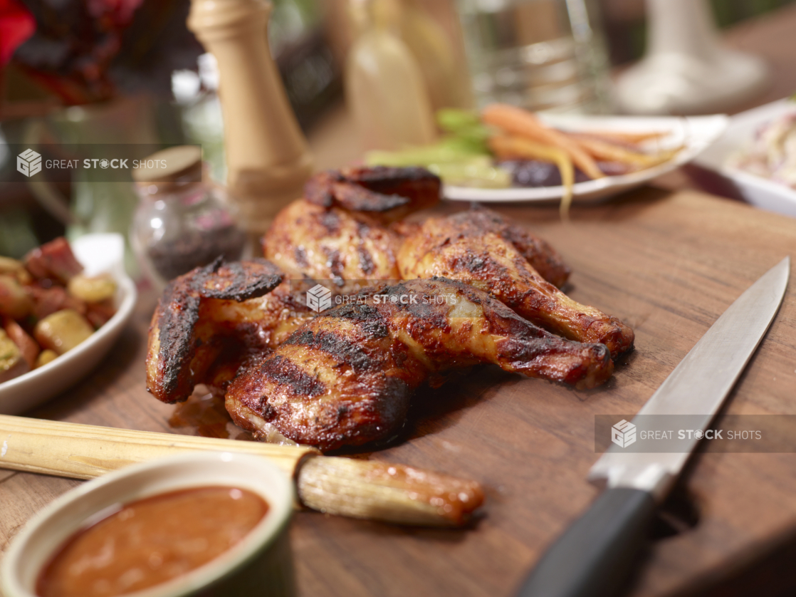 Barbecued butterflied chicken on a wood cutting board beside a dish of BBQ sauce and sauce brush, other dishes in background