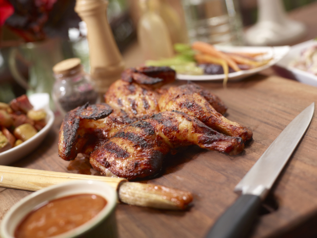 Barbecued butterflied chicken on a wood cutting board beside a dish of BBQ sauce and sauce brush, other dishes in background