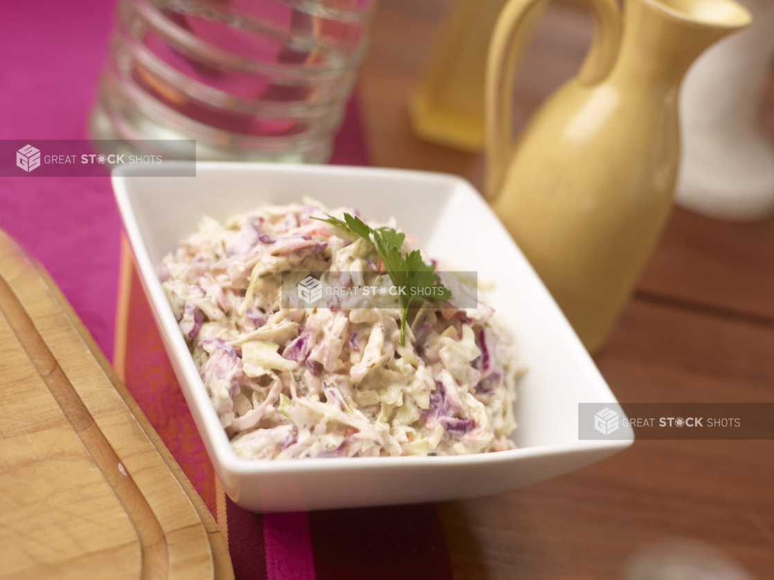 Coleslaw garnished with a sprig of fresh parsley in a square white dish on a wood table with pink table runner, yellow jug in background