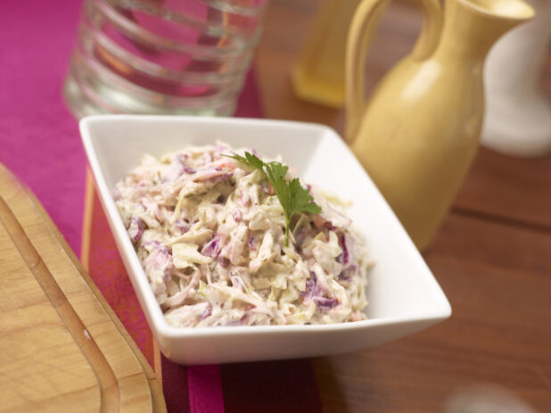 Coleslaw garnished with a sprig of fresh parsley in a square white dish on a wood table with pink table runner, yellow jug in background