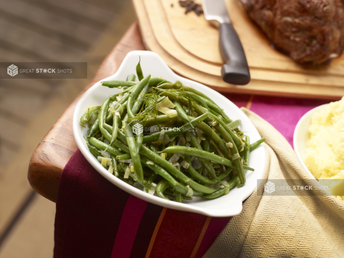 Green beans with leeks and fennel in a white serving dish, roast and mashed potatoes in background