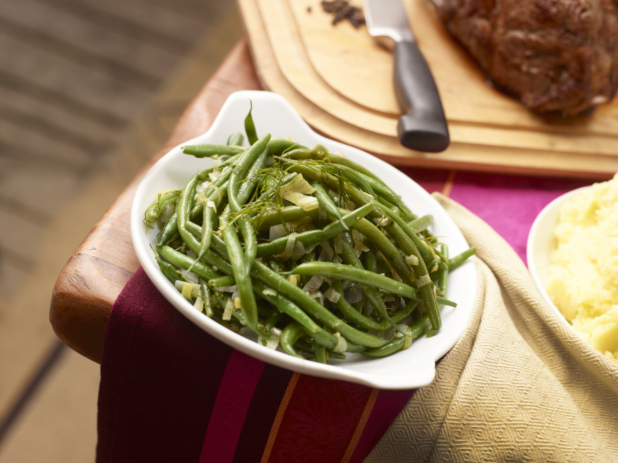 Green beans with leeks and fennel in a white serving dish, roast and mashed potatoes in background