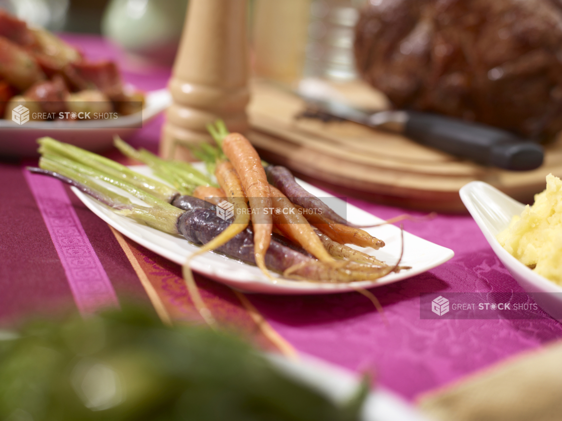 Whole orange and purple heirloom carrots with green tops on a white plate on a buffet table with a pink tablecloth, other buffet items surrounding