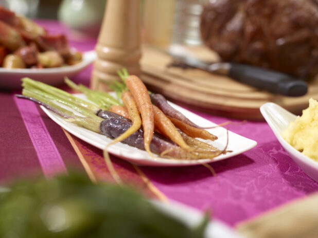 Whole orange and purple heirloom carrots with green tops on a white plate on a buffet table with a pink tablecloth, other buffet items surrounding