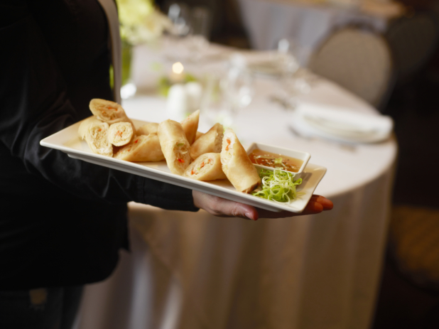 Hand holding a small platter of halved spring rolls, green onion garnish and dipping sauce, catered event setting