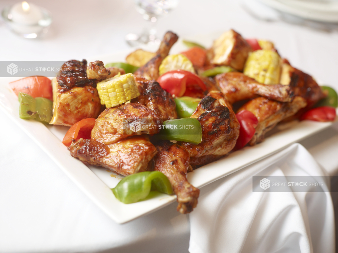 Barbecued chicken legs with roasted peppers and chunks of corn on the cob on a white platter, white table setting