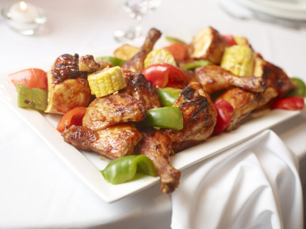 Barbecued chicken legs with roasted peppers and chunks of corn on the cob on a white platter, white table setting