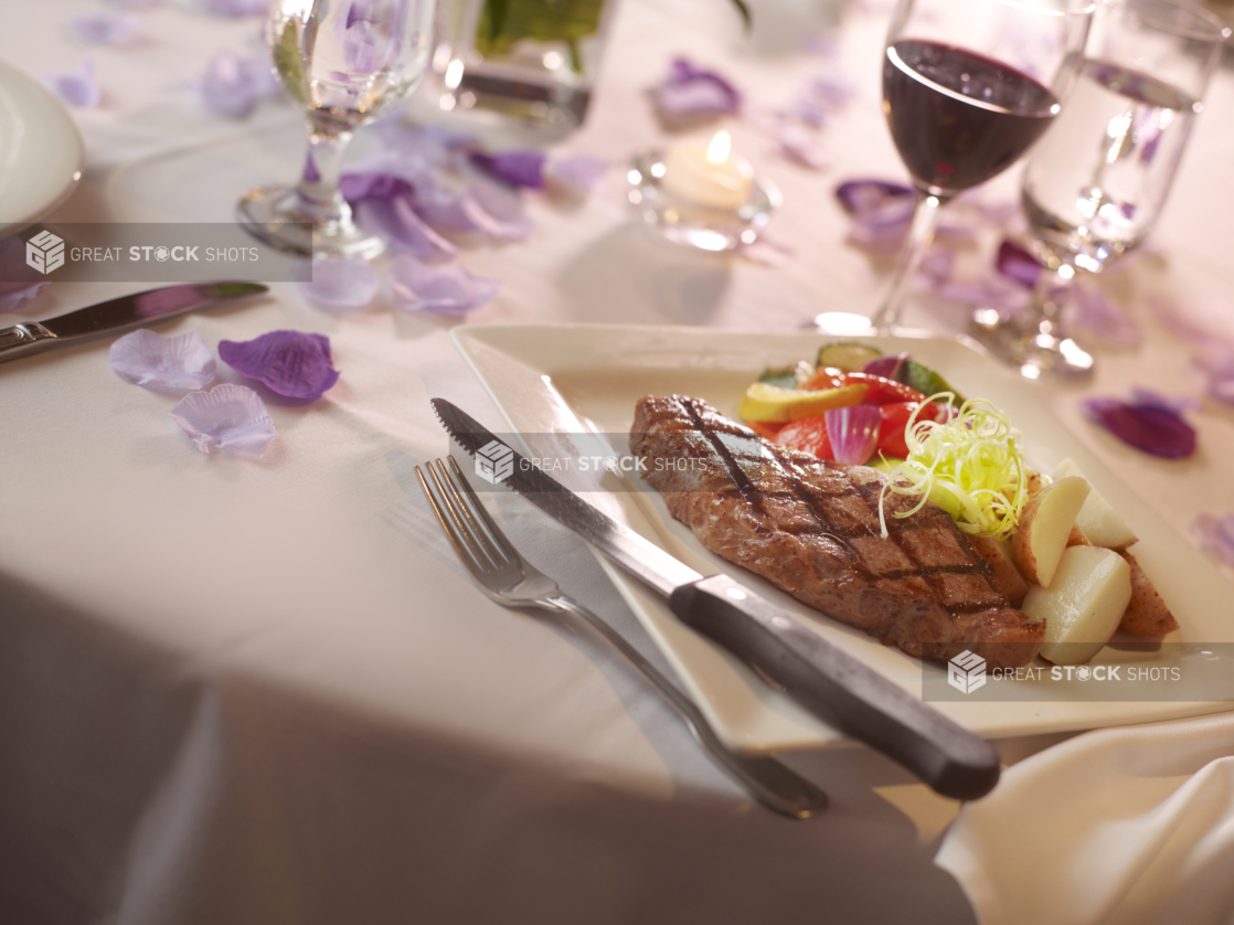 Steak dinner with potatoes and vegetables and a glass of red wine in a white table setting, purple flower petals surrounding