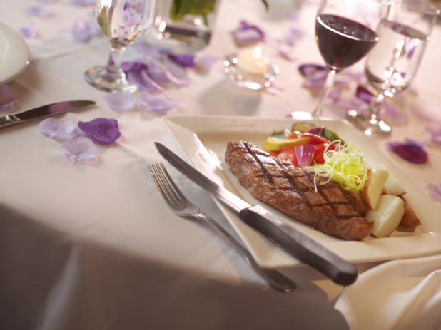 Steak dinner with potatoes and vegetables and a glass of red wine in a white table setting, purple flower petals surrounding
