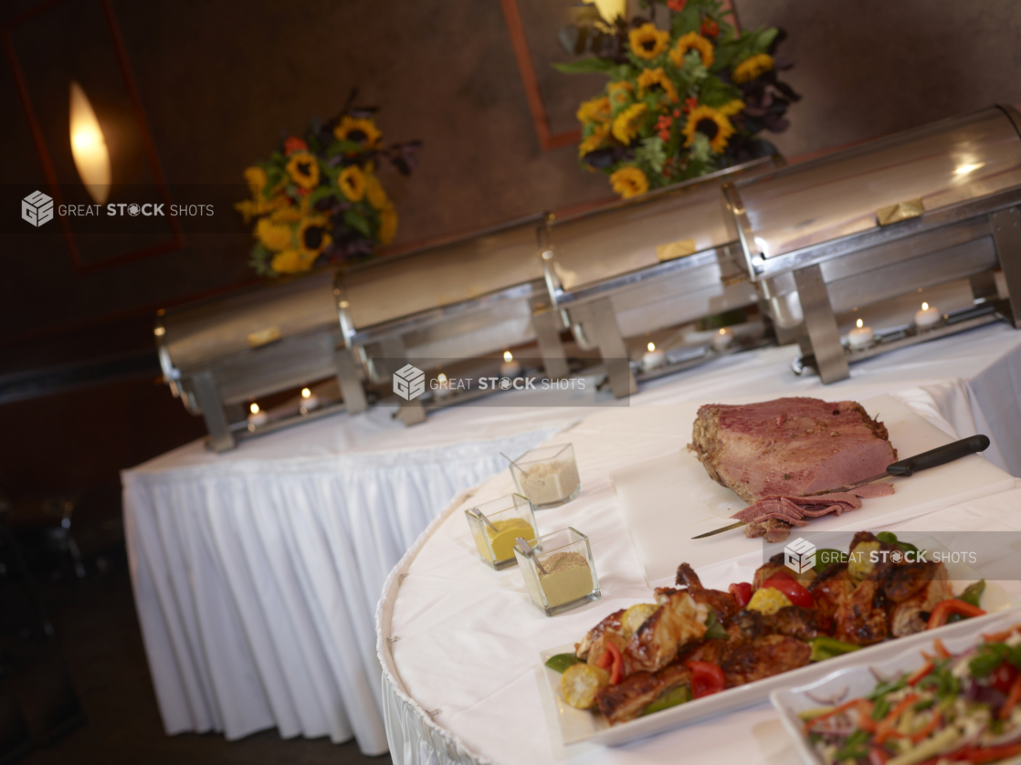 A white buffet table with a row of steamers and a corned beef, partially sliced, in foreground