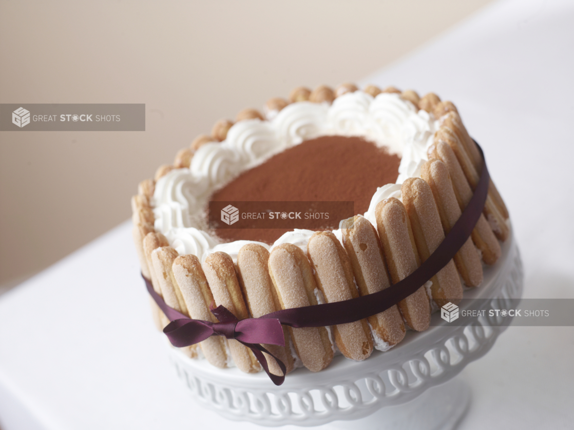 tiramisu cake with a purple ribbon finger cookies on a white cake stand and table cloth