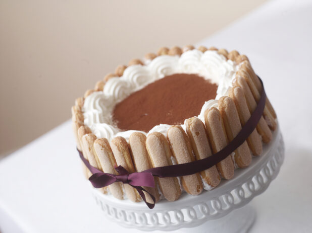 tiramisu cake with a purple ribbon finger cookies on a white cake stand and table cloth