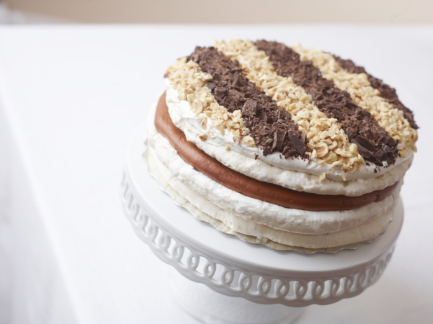 layered caramel crunch cake topped with chopped peanuts almonds and chocolate on a white cake stand