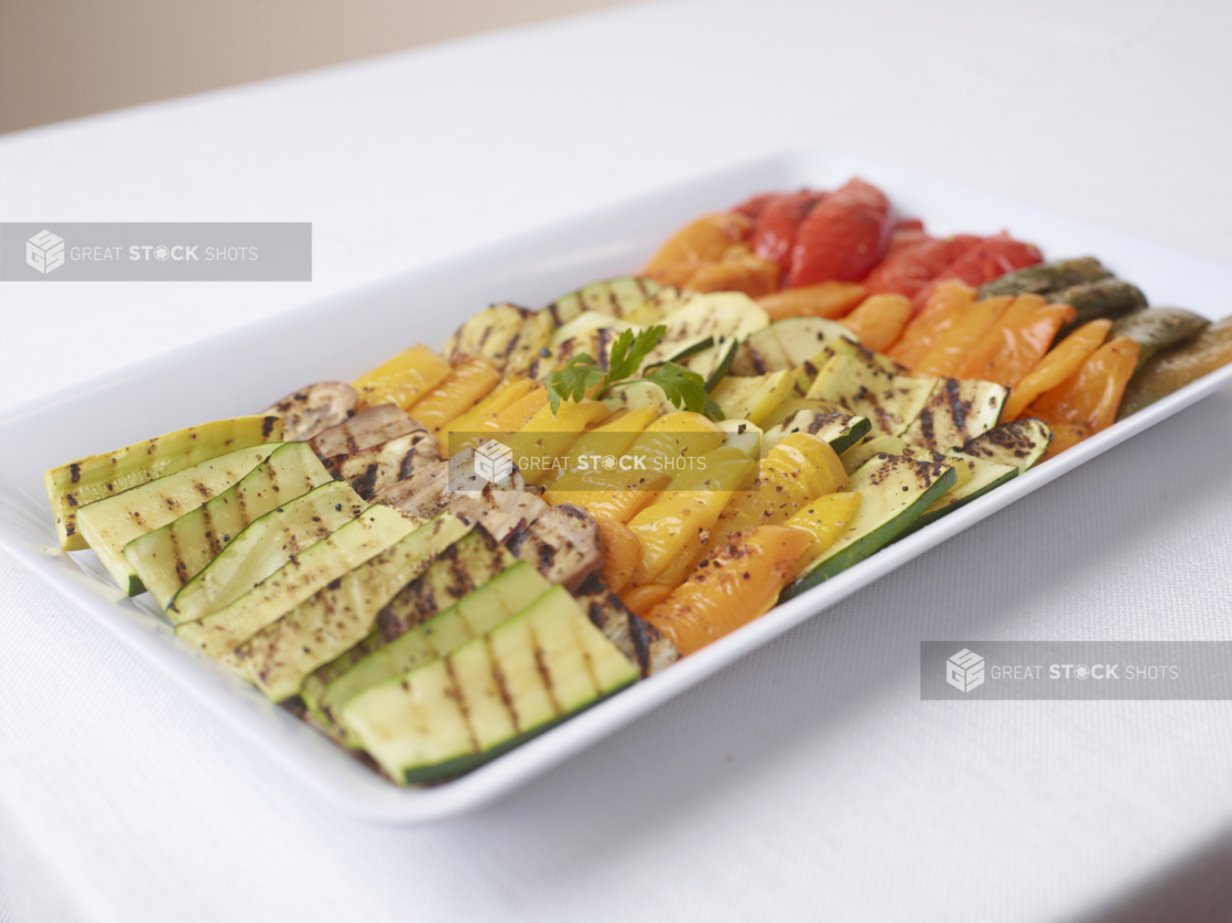 white tray of grilled zucchini eggplant yellow orange red peppers on white table cloth