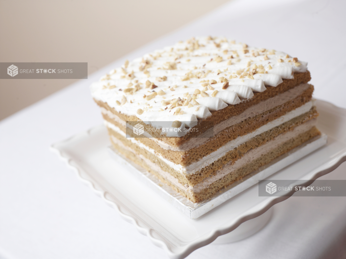 square 4 layered cake topped with whipped cream and chopped almonds on a white cake stand