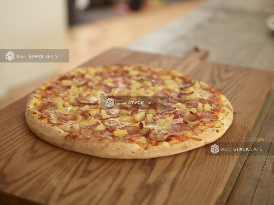 Whole Hawaiian Pizza with Pineapples, Bacon and Sliced Ham on a Wooden Cutting Board in a Dining Room Setting