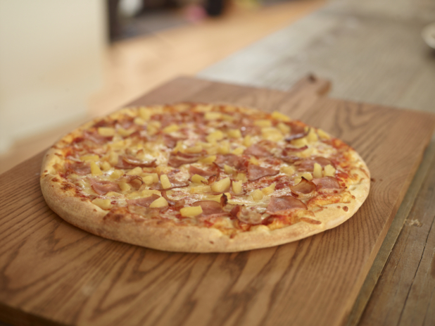 Whole Hawaiian Pizza with Pineapples, Bacon and Sliced Ham on a Wooden Cutting Board in a Dining Room Setting