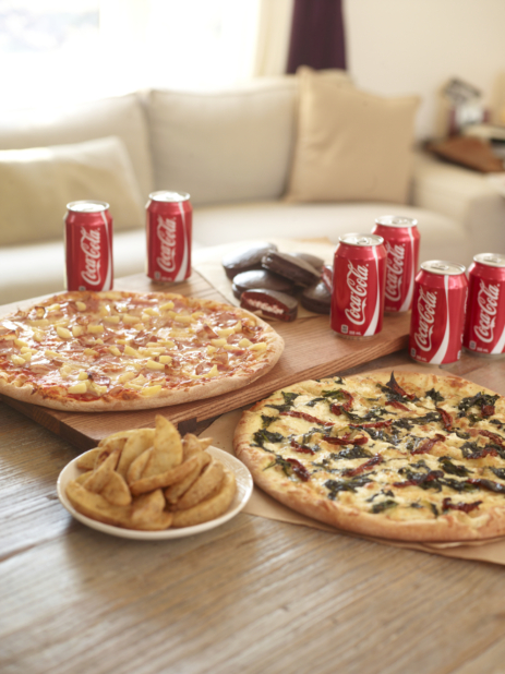 Pizza Meal - Hawaiian pizza, vegetarian pizza, potato wedges, cans of cola, and chocolate snack cakes on a wood table