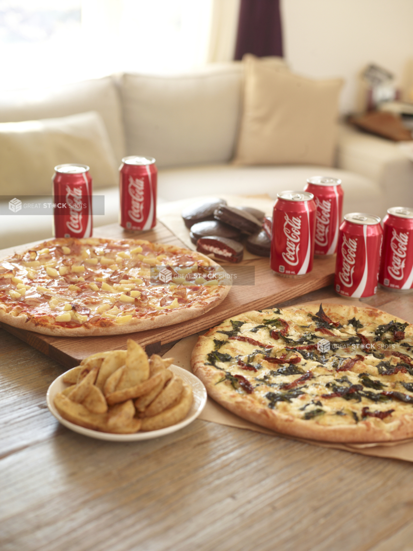 Pizza Meal - Hawaiian pizza, vegetarian pizza, potato wedges, cans of cola, and chocolate snack cakes on a wood table