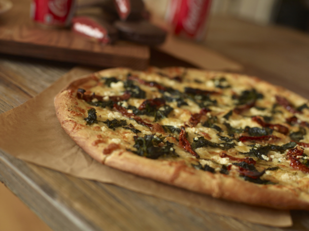 Whole unsliced spinach, sundried tomato and goat cheese pizza on parchment on a wood tabletop