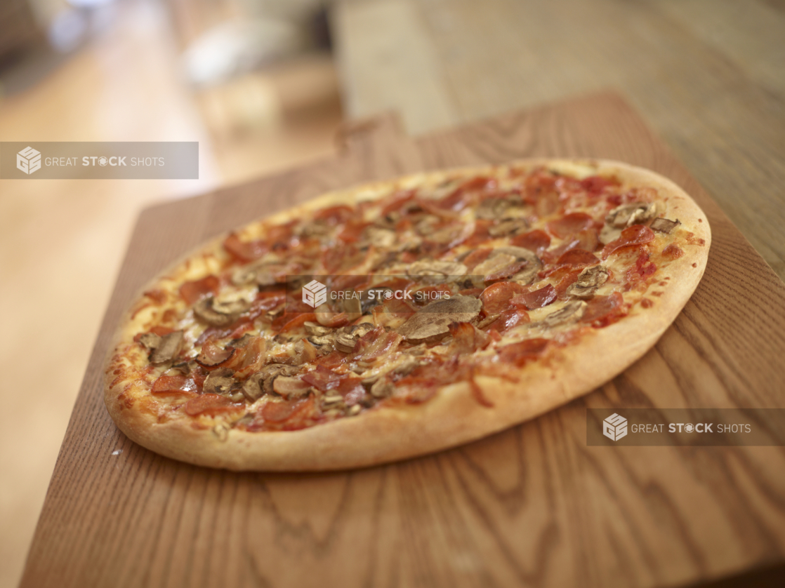 Whole Canadian Pizza with Pepperoni, Bacon and Mushrooms on a Wooden Cutting Board in a Dining Room Setting