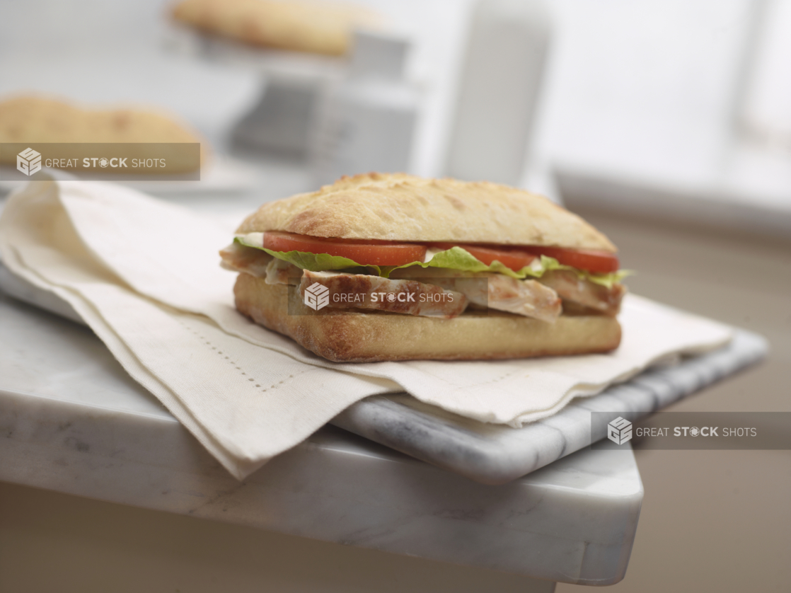 Grilled Chicken Sandwich with Lettuce and Tomatoes on a Ciabatta Bun on a White Cloth and Marble Surface in an Indoor Setting