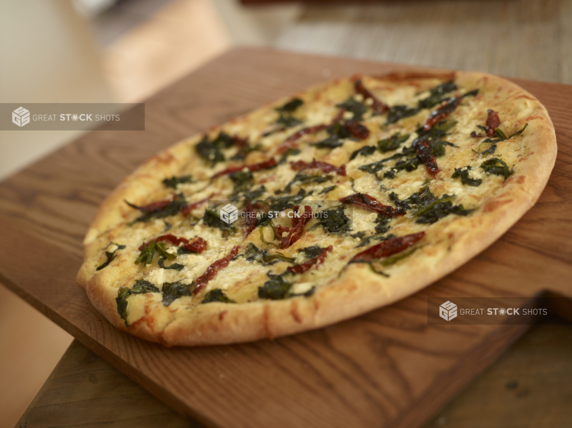 Whole 3-Topping Pizza with Sautéed Spinach, Sun-dried Tomatoes and Feta Cheese on a Wooden Cutting Board in a Dining Room Setting