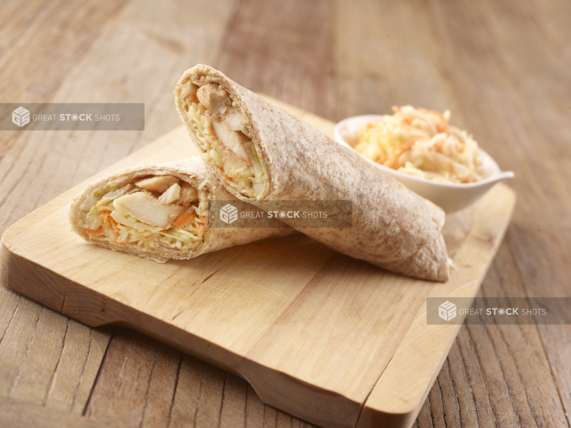 Fried Fish and Shredded Vegetables in a Whole Wheat Wrap Cut in Half on a Wooden Cutting Board with a Side Dish of Coleslaw on a Rustic Wooden Surface