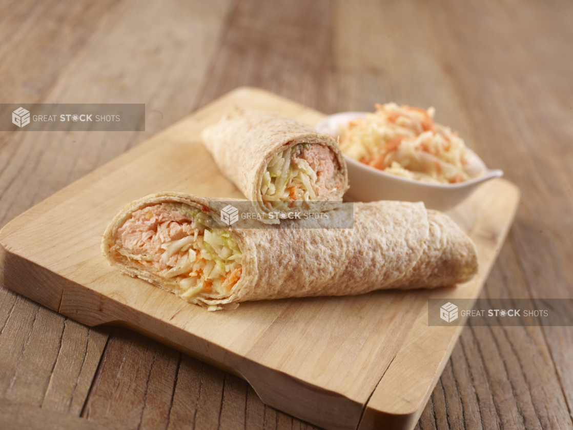 Salmon and Shredded Vegetables in a Whole Wheat Wrap Cut in Half on a Wooden Cutting Board with a Side Dish of Coleslaw on a Rustic Wooden Surface