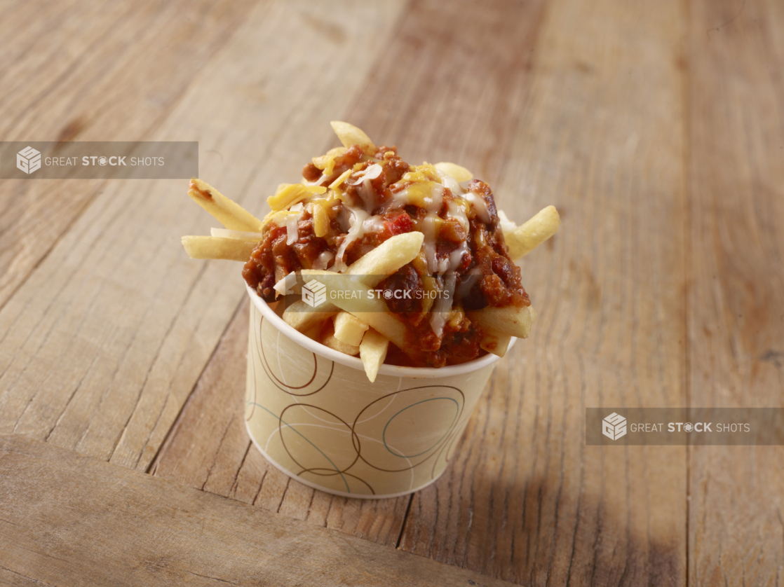 Thick Cut French Fries Topped with Chilli Cheese in a Paper Take-Out Bowl on a Weathered Wooden Surface in an Indoor Setting