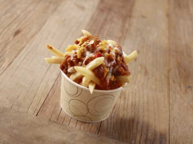 Thick Cut French Fries Topped with Chilli Cheese in a Paper Take-Out Bowl on a Weathered Wooden Surface in an Indoor Setting