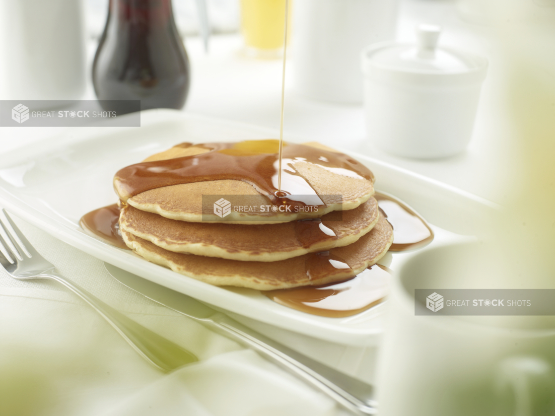 A Platter with a Stack of Buttermilk Pancakes with Maple Syrup Being Poured Overtop, in a Restaurant Setting