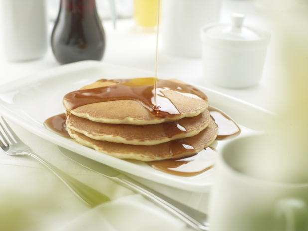 A Platter with a Stack of Buttermilk Pancakes with Maple Syrup Being Poured Overtop, in a Restaurant Setting
